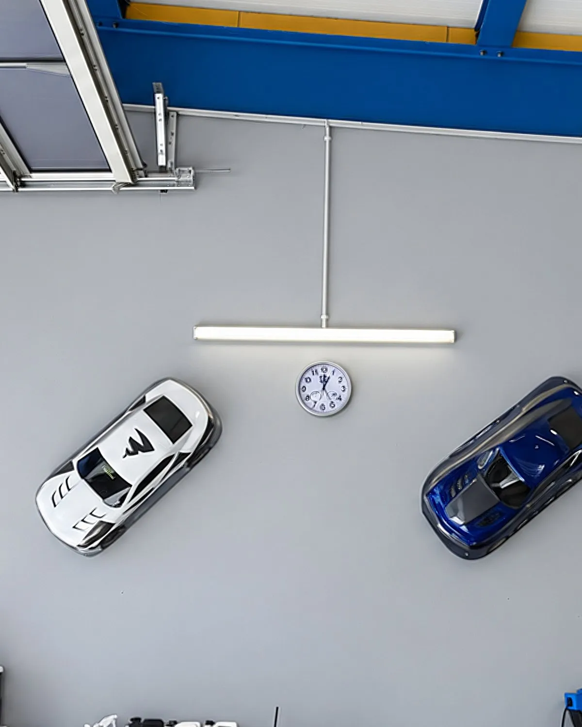 Hallenwand mit zwei Automodellen, Uhr und Lichtleiste unter blauen Stahlträgern