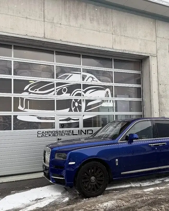 Blauer Rolls-Royce Cullinan vor dem Werkstatttor mit dem Lind-Schriftzug, Schnee am Boden